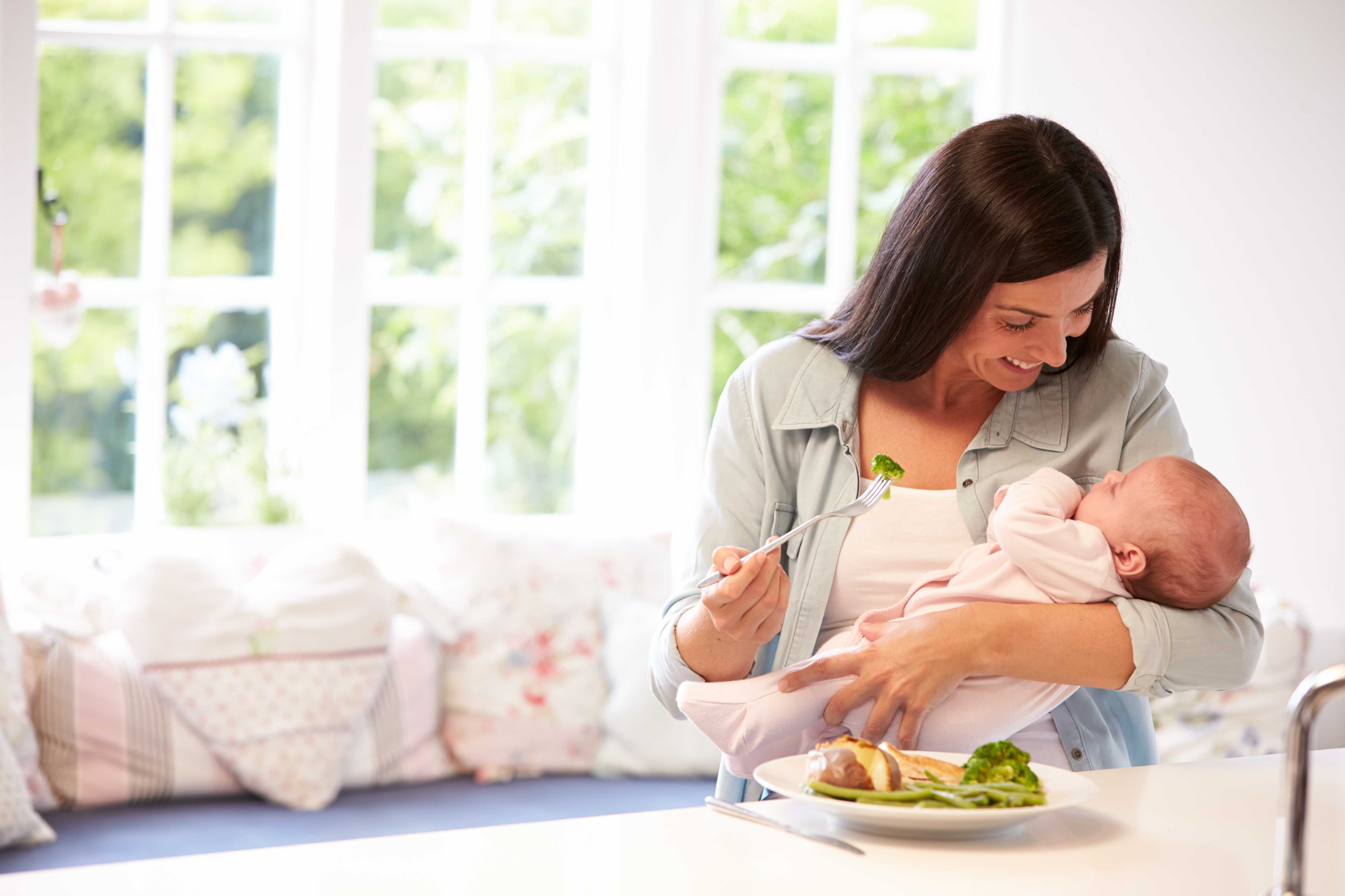 Mother feeding herself while holding a baby, healthy meal with broccoli.