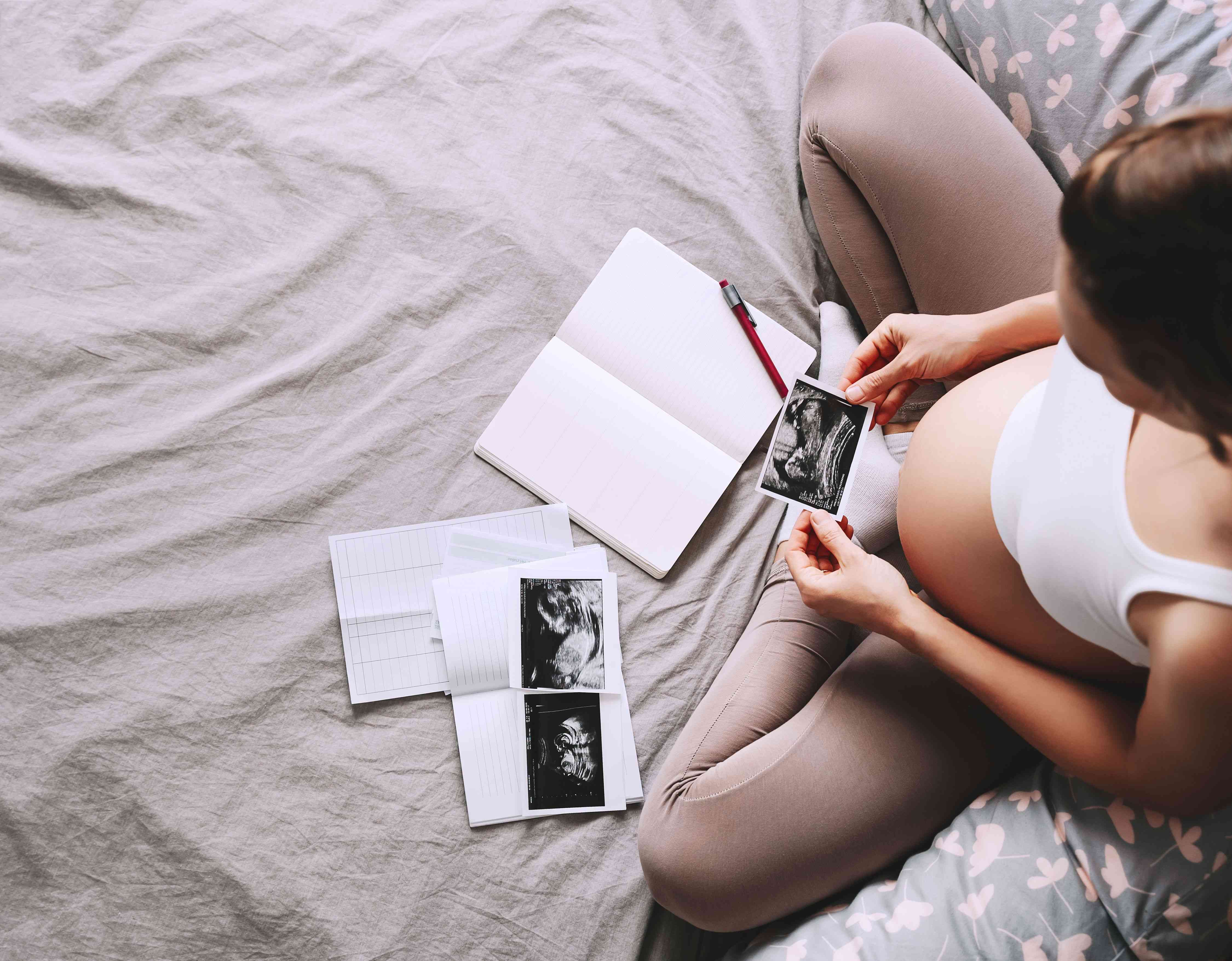 Pregnant woman holding an ultrasound image while sitting with an open journal and medical reports on a bed.