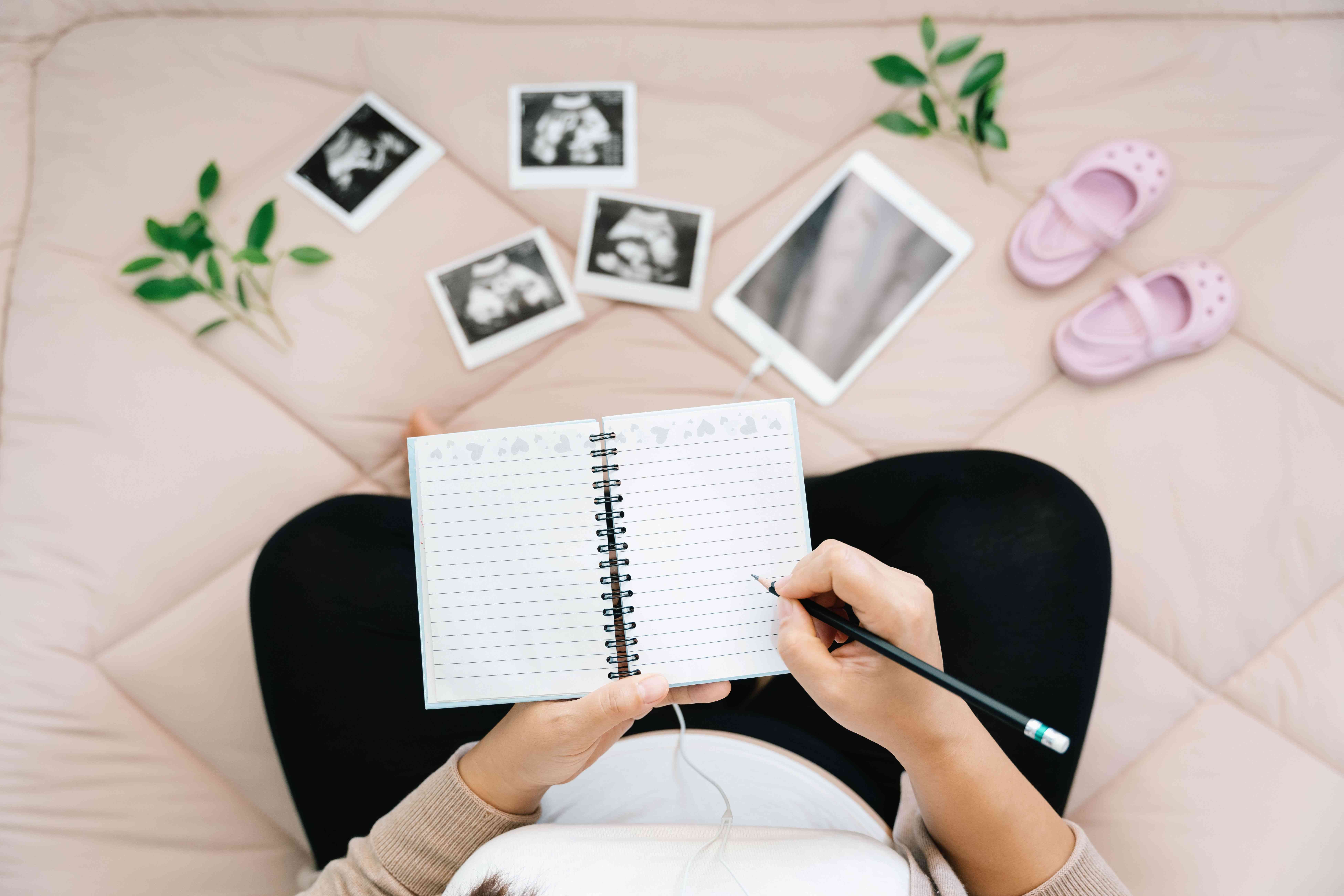 Top view of a pregnant woman writing in a pregnancy journal surrounded by ultrasound photos, a tablet, and baby girl shoes.