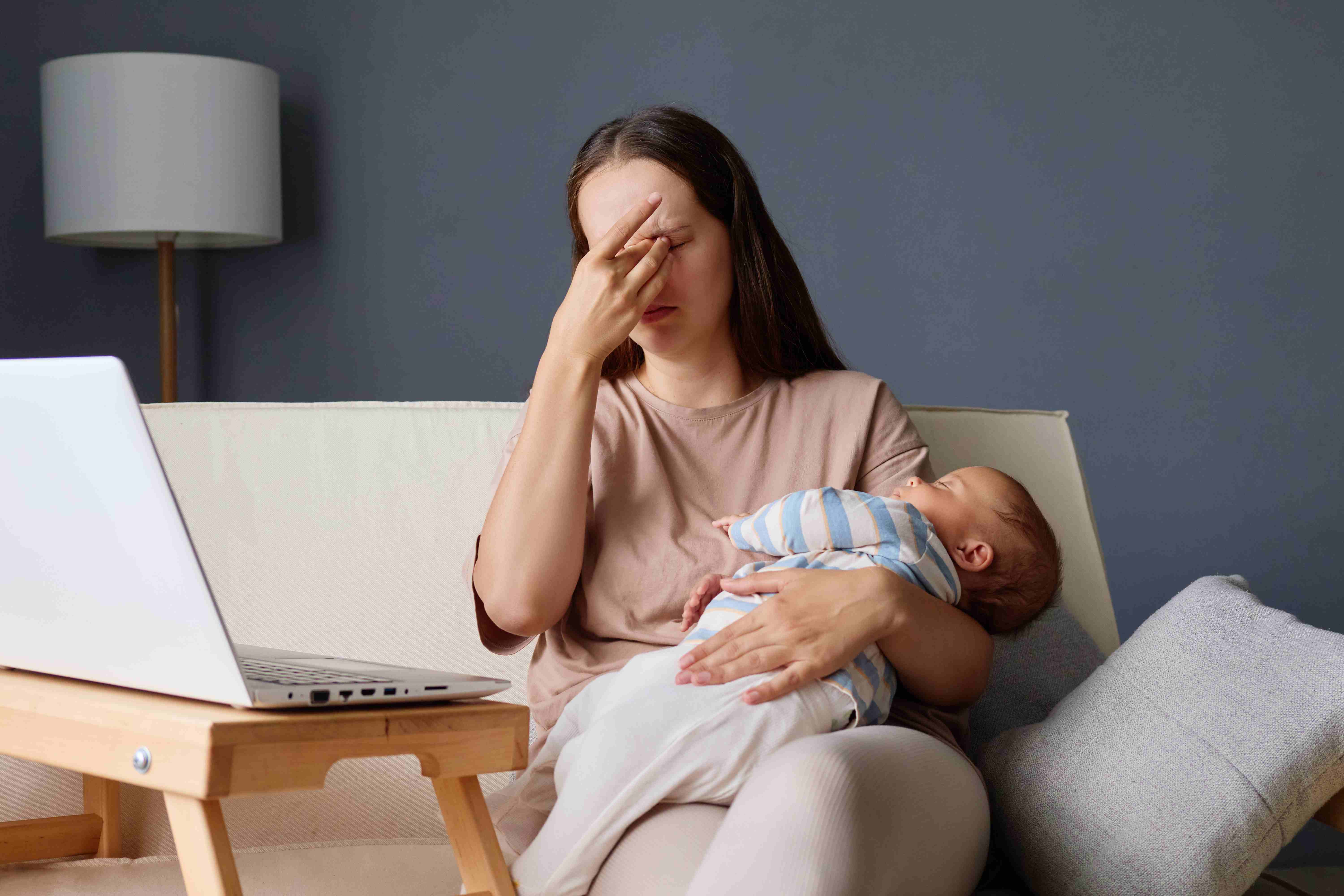Tired mother holding a newborn while working on a laptop at home, rubbing her eyes from exhaustion.