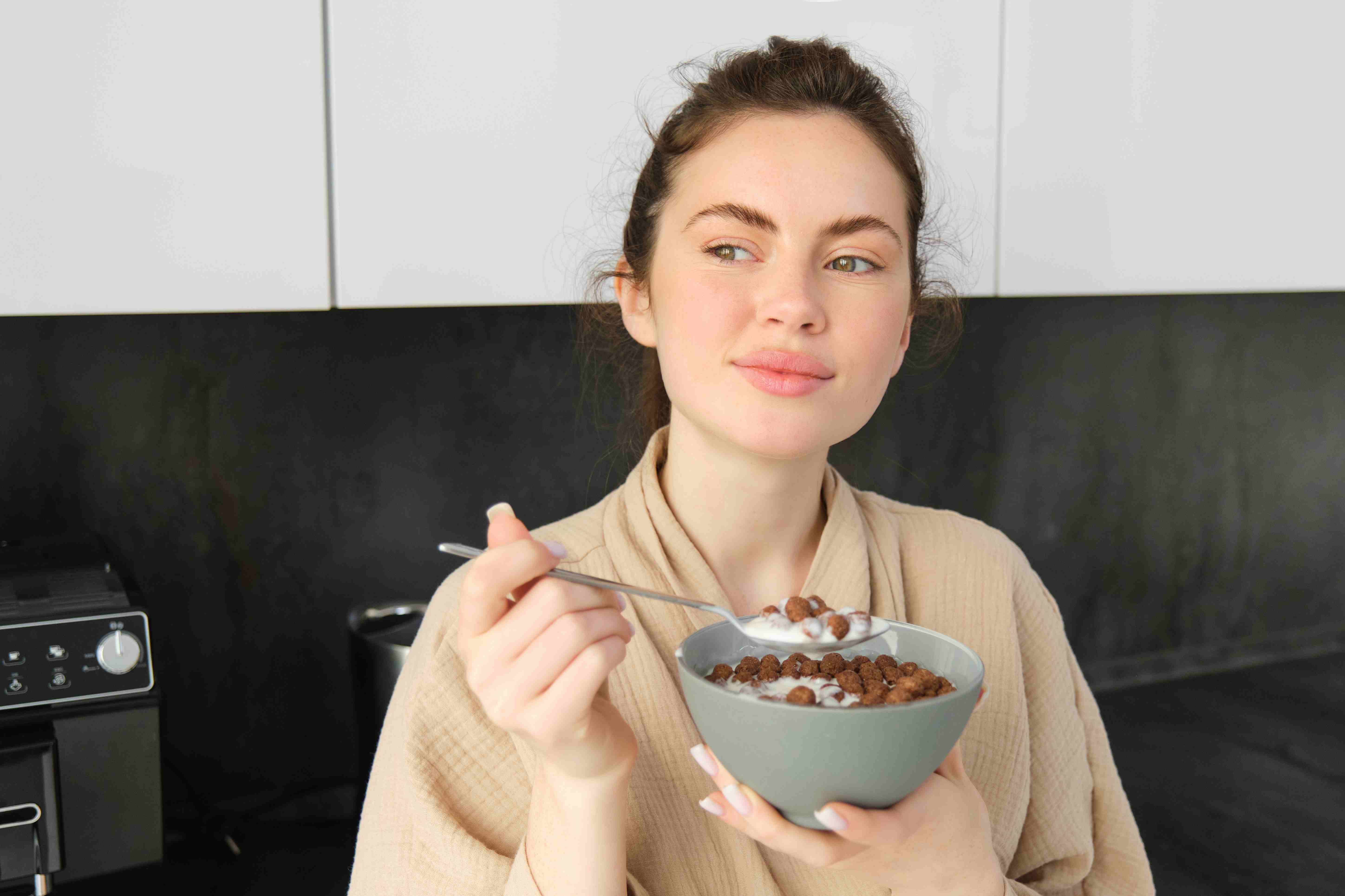 Relaxed woman in a robe eating a bowl of cereal in the kitchen, reflecting a mindful moment in postpartum eating habits.