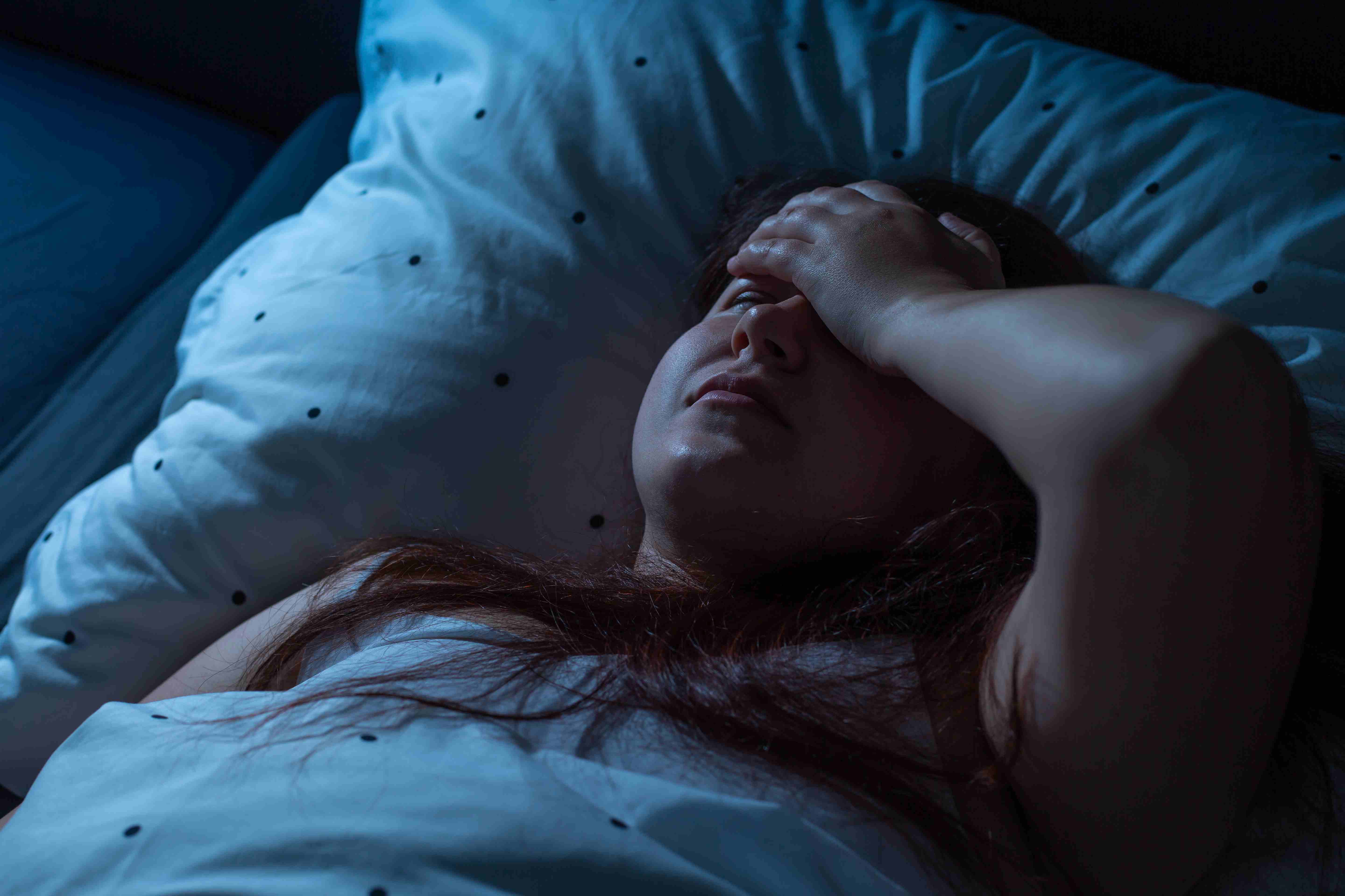 Woman lying awake in bed at night, looking distressed with hand on forehead due to Postpartum Night Sweats.