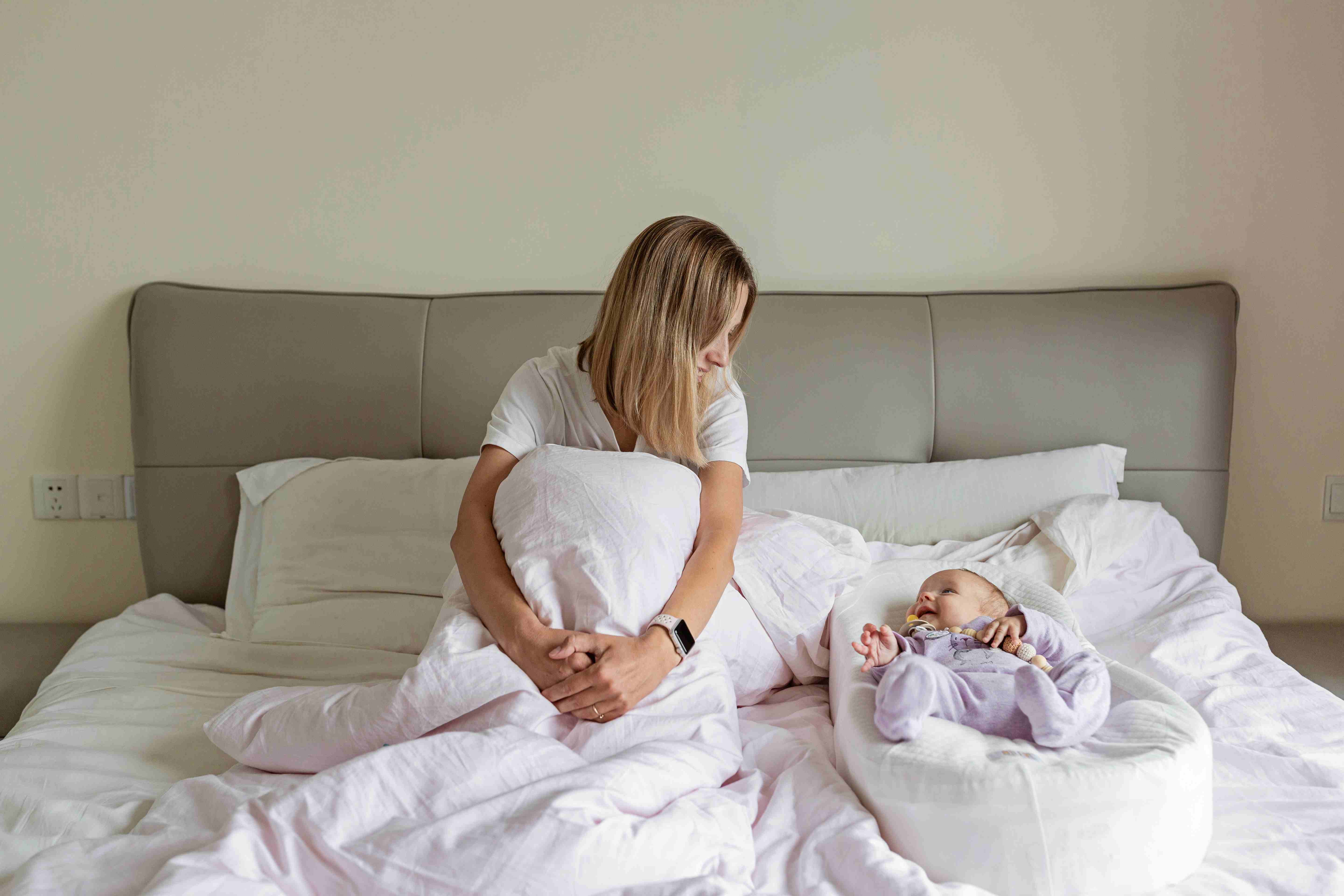 Tired mother sitting up in bed while her newborn sleeps in a bassinet nearby.