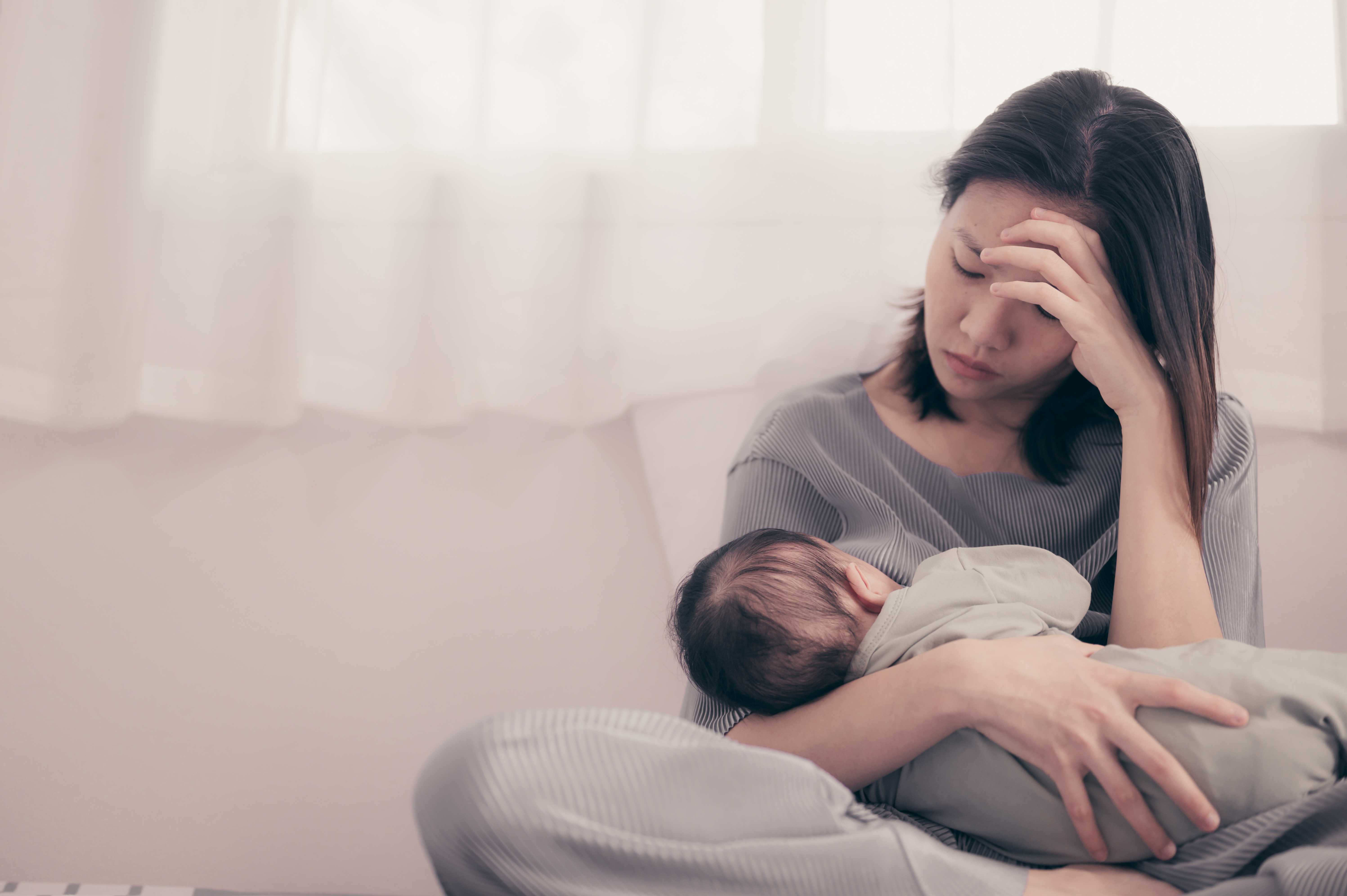 Tired mother holding her baby while breastfeeding, sitting by a window with her hand on her forehead, showing signs of postnatal exhaustion or depression.