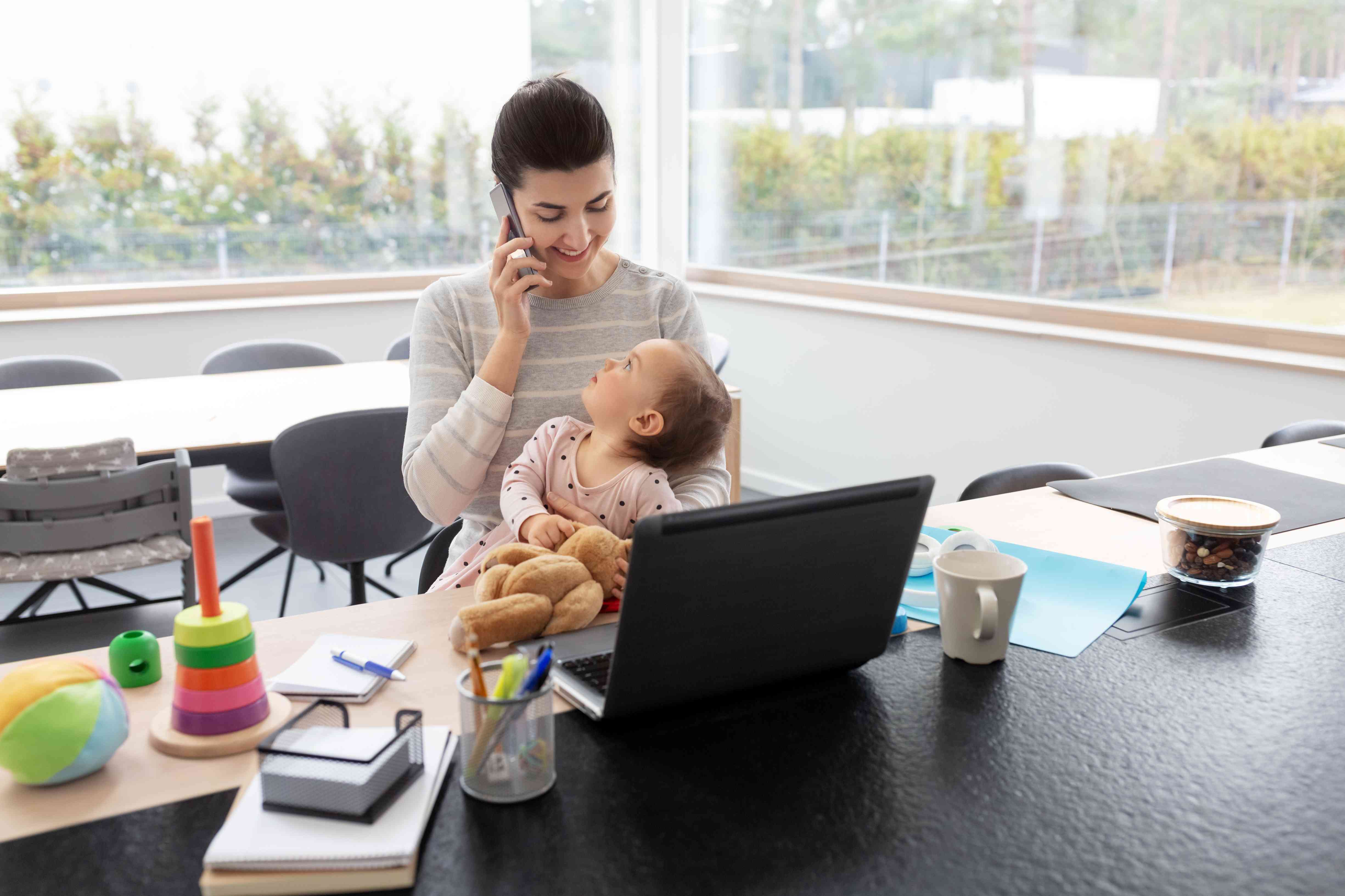  A mother holding her baby while working on a laptop in a home office setup.