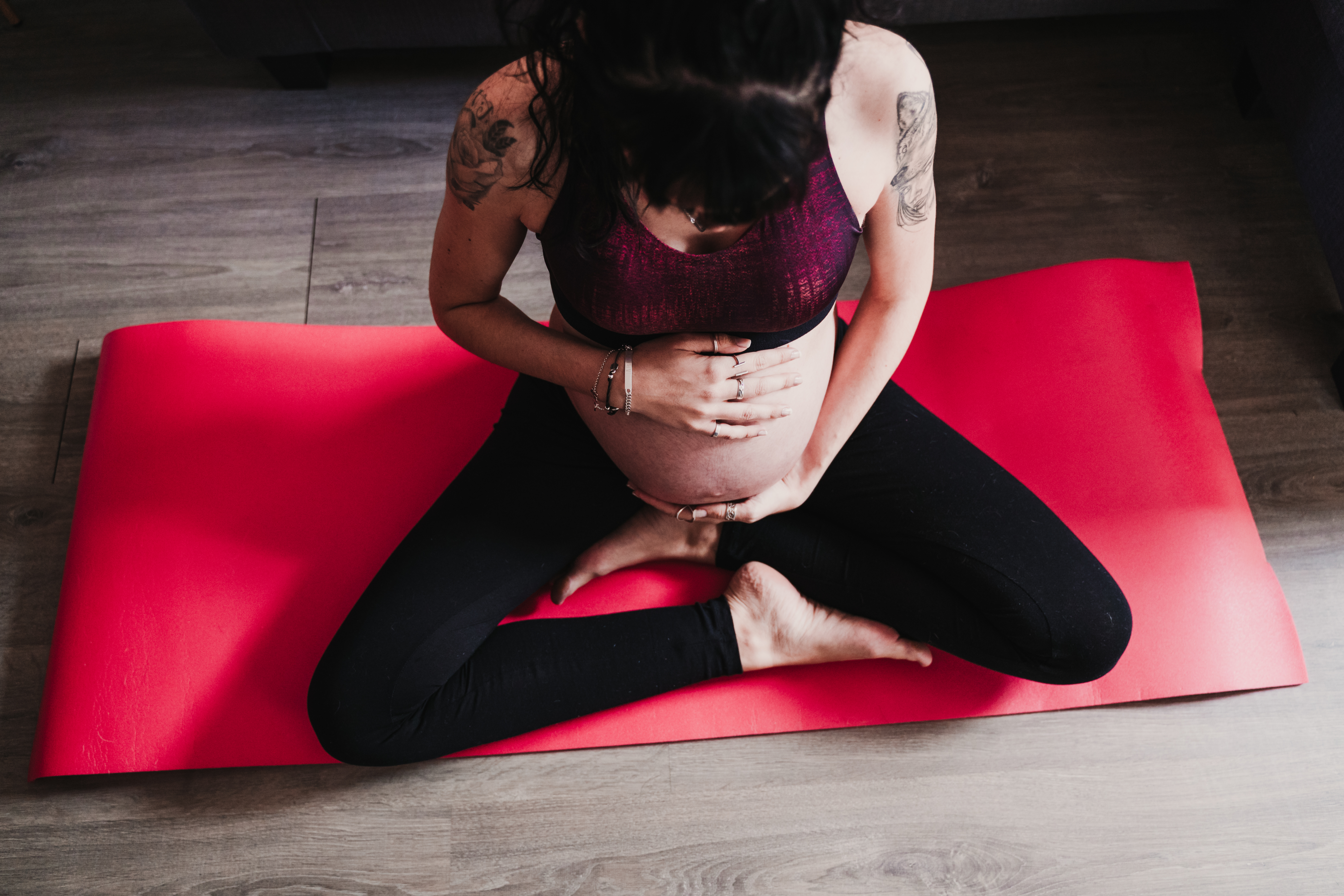 Pregnant woman holding her belly in a seated butterfly pose on a red yoga mat at home.
